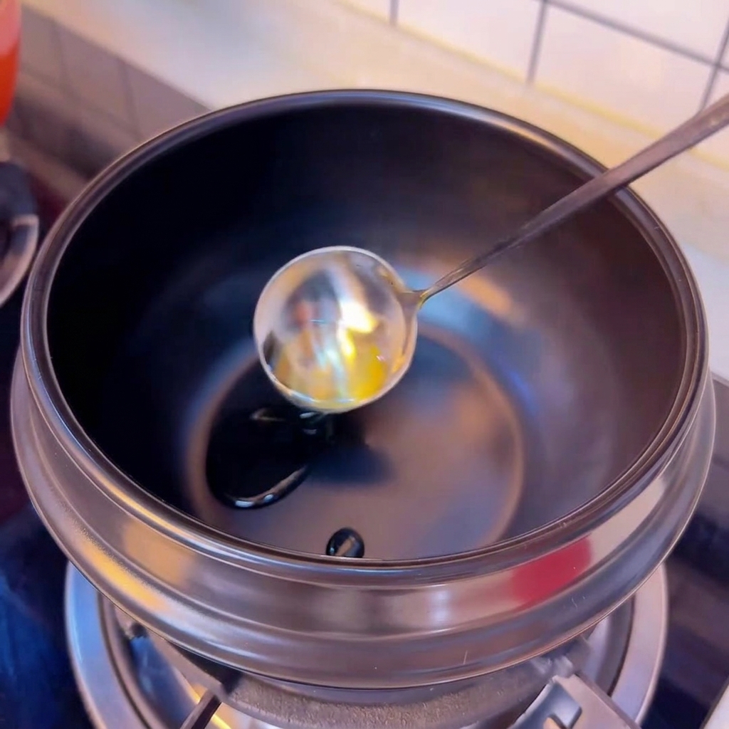 Camellia oil being poured from a silver ladle into a preheated black clay cooking pot.