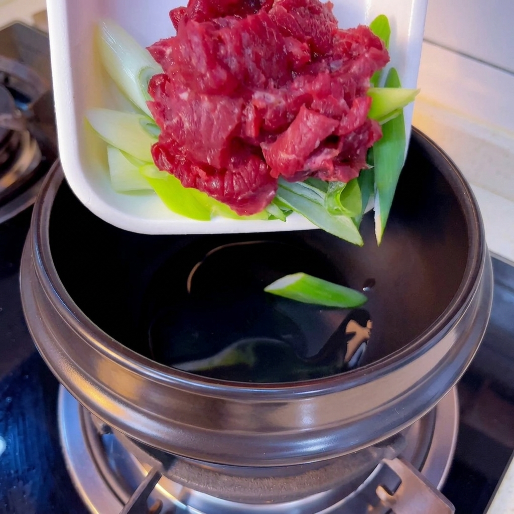 Raw red beef slices and chopped green onions being added into a black clay pot from a white tray.