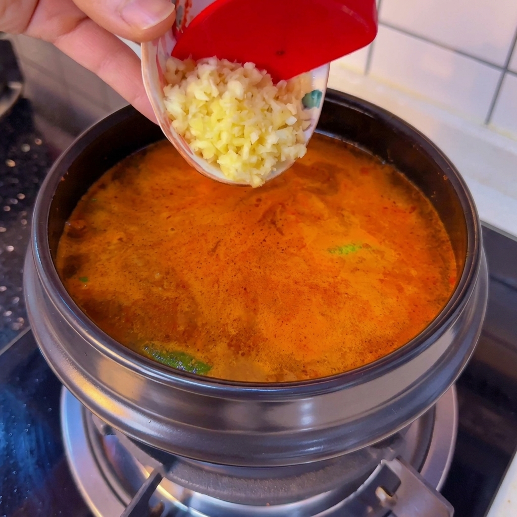 Minced garlic being scraped from a small patterned bowl into a bubbling pot of spicy Korean soft tofu stew.