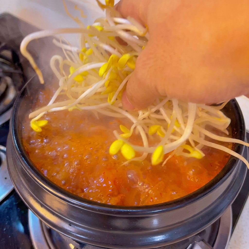 A hand placing a bundle of fresh yellow bean sprouts into a bubbling, orange-red soup pot.