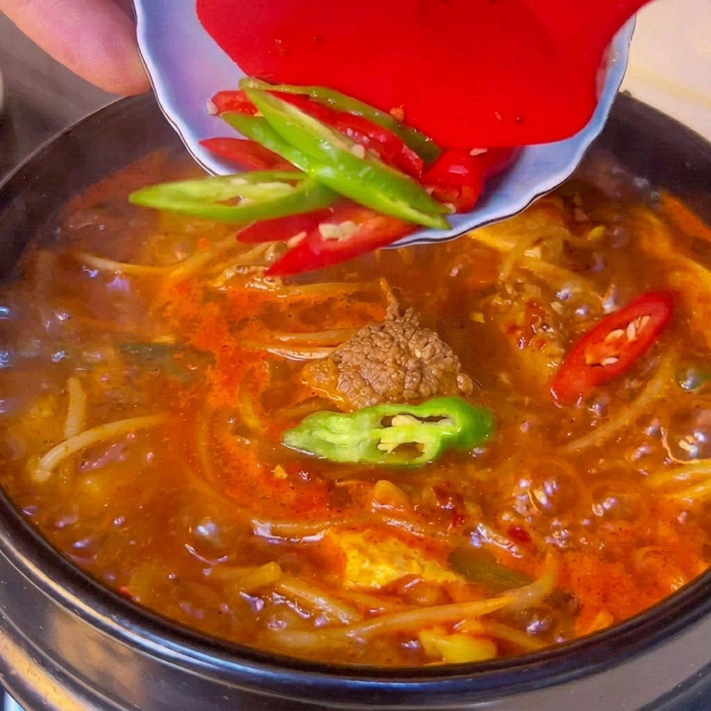 Fresh green and red chili slices being scraped from a small white bowl onto the surface of a boiling spicy tofu stew.