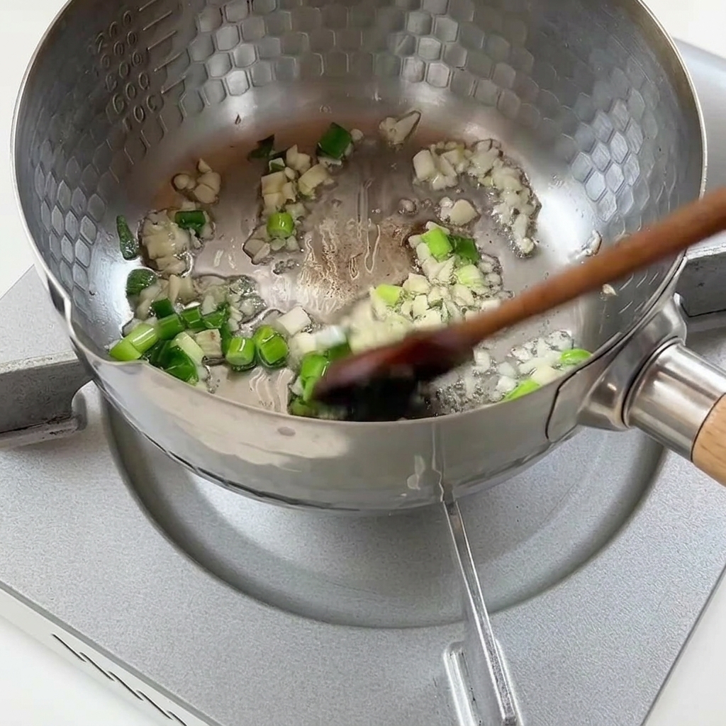 Chopped green onions and minced garlic sautéing in hot oil inside a textured metal pot.