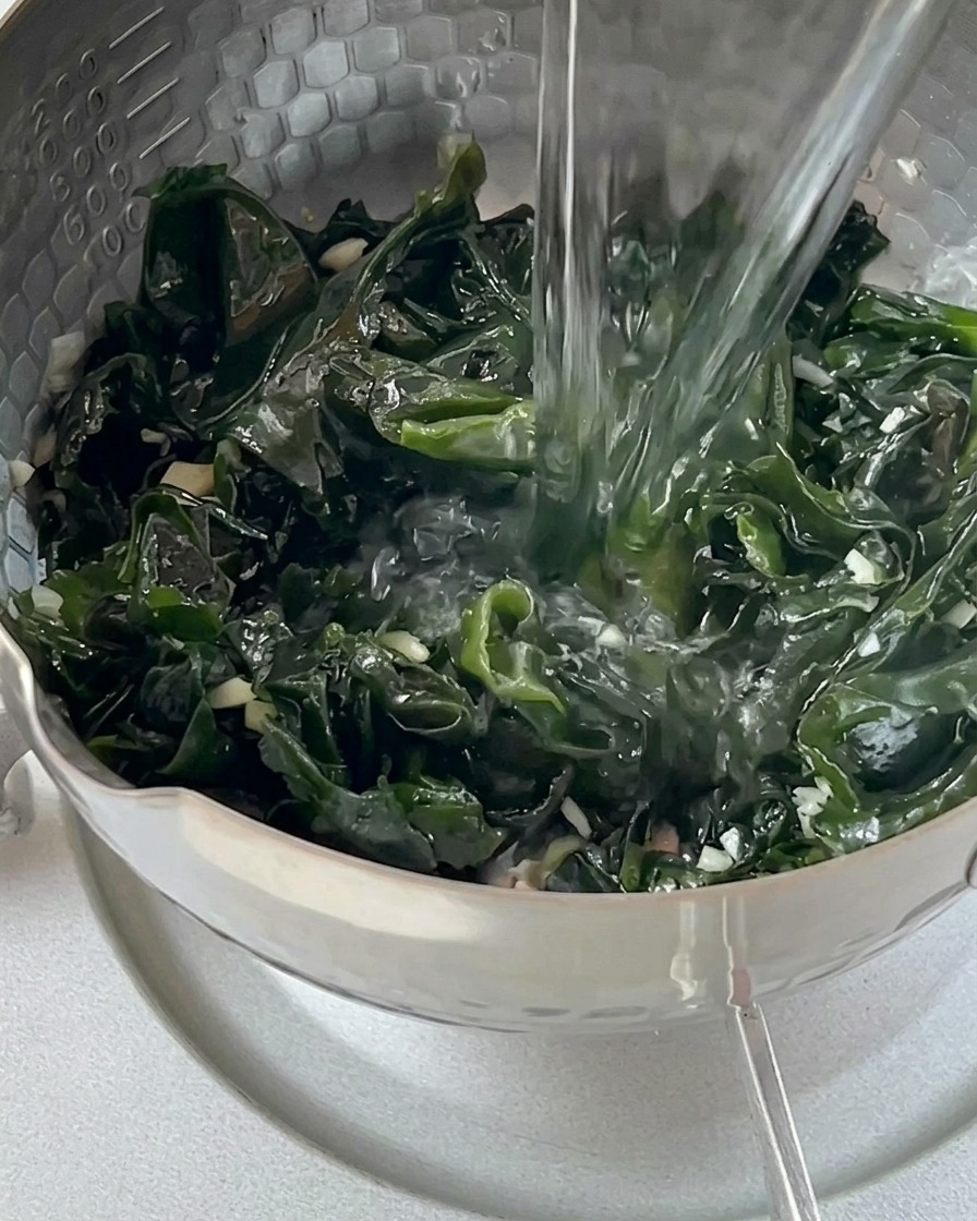 Boiling water pouring from a glass kettle over green wakame in a pot.
