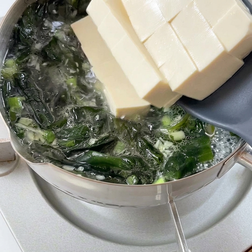 Thick slices of soft white tofu sliding from a gray spatula into a bubbling pot of wakame broth.