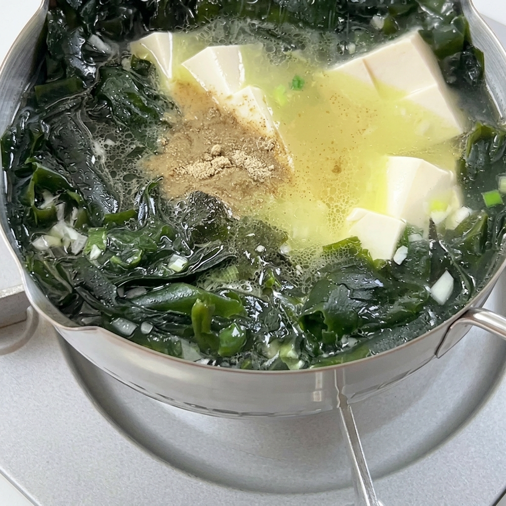 Pepper powder being sprinkled into a bubbling pot of soup containing wakame seaweed and cubed tofu.