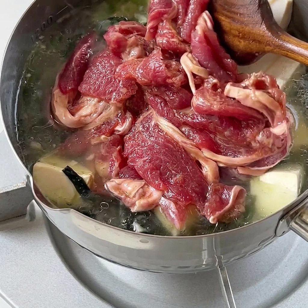 Marinated raw beef slices being gently added and stirred into a hot pot of wakame and tofu soup.