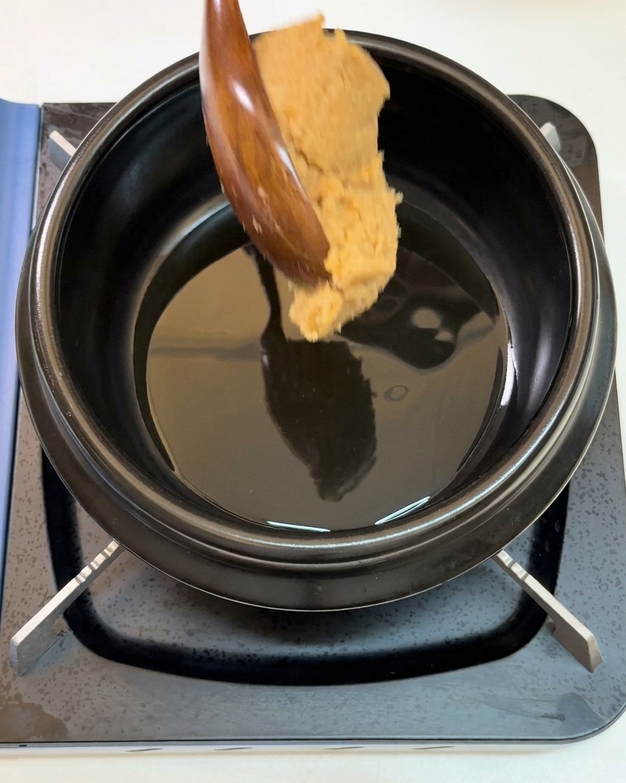 A large spoonful of thick soybean paste being lowered from a wooden spoon into a black pot filled with shimmering sesame oil.