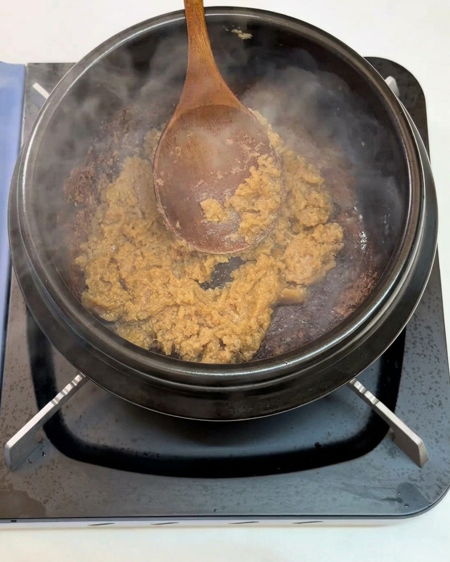 A wooden spoon stir-frying minced garlic and soybean paste inside a heated black earthenware pot on a stove.