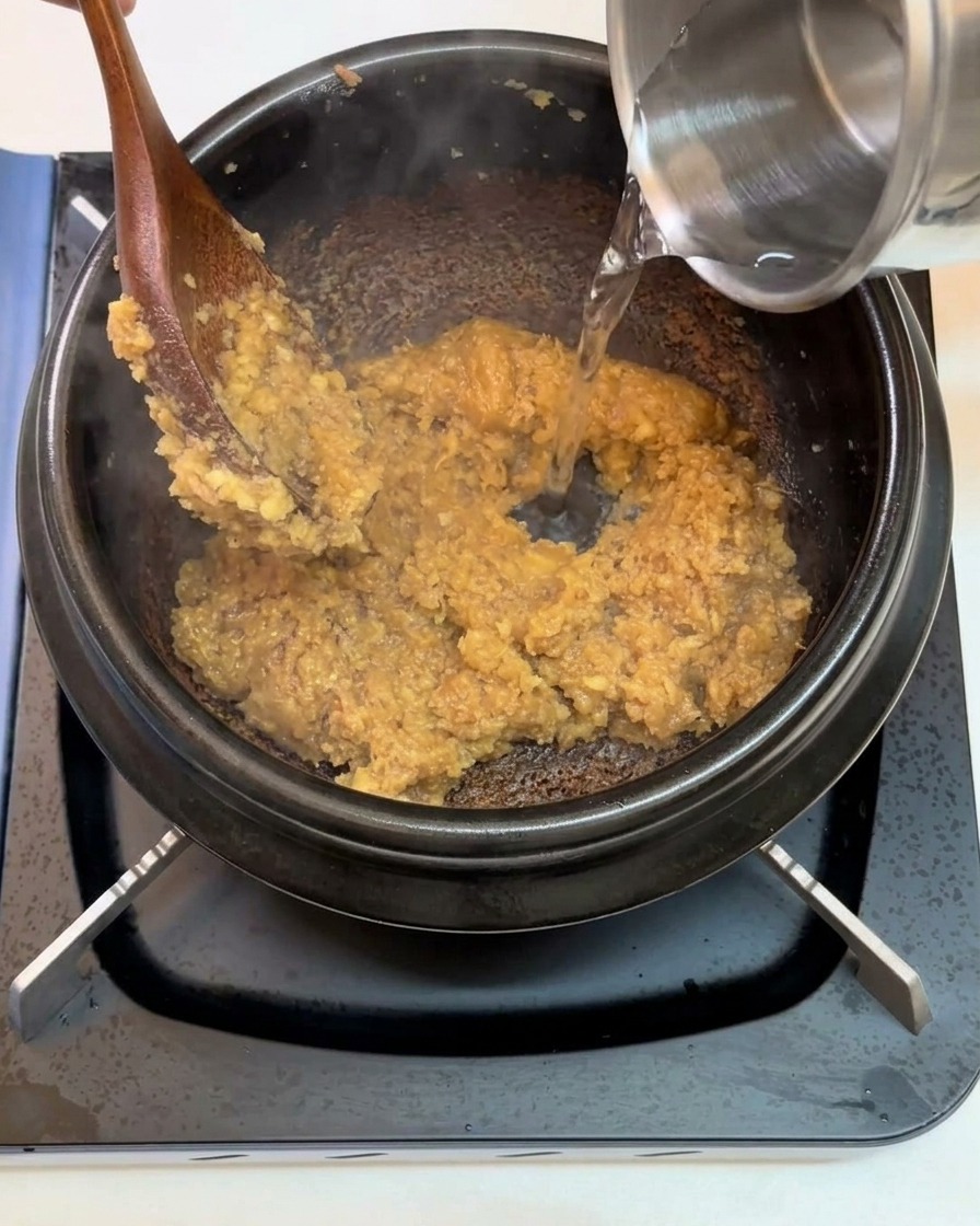 Water being poured from a metallic container into a black pot containing the stir-fried soybean paste base.