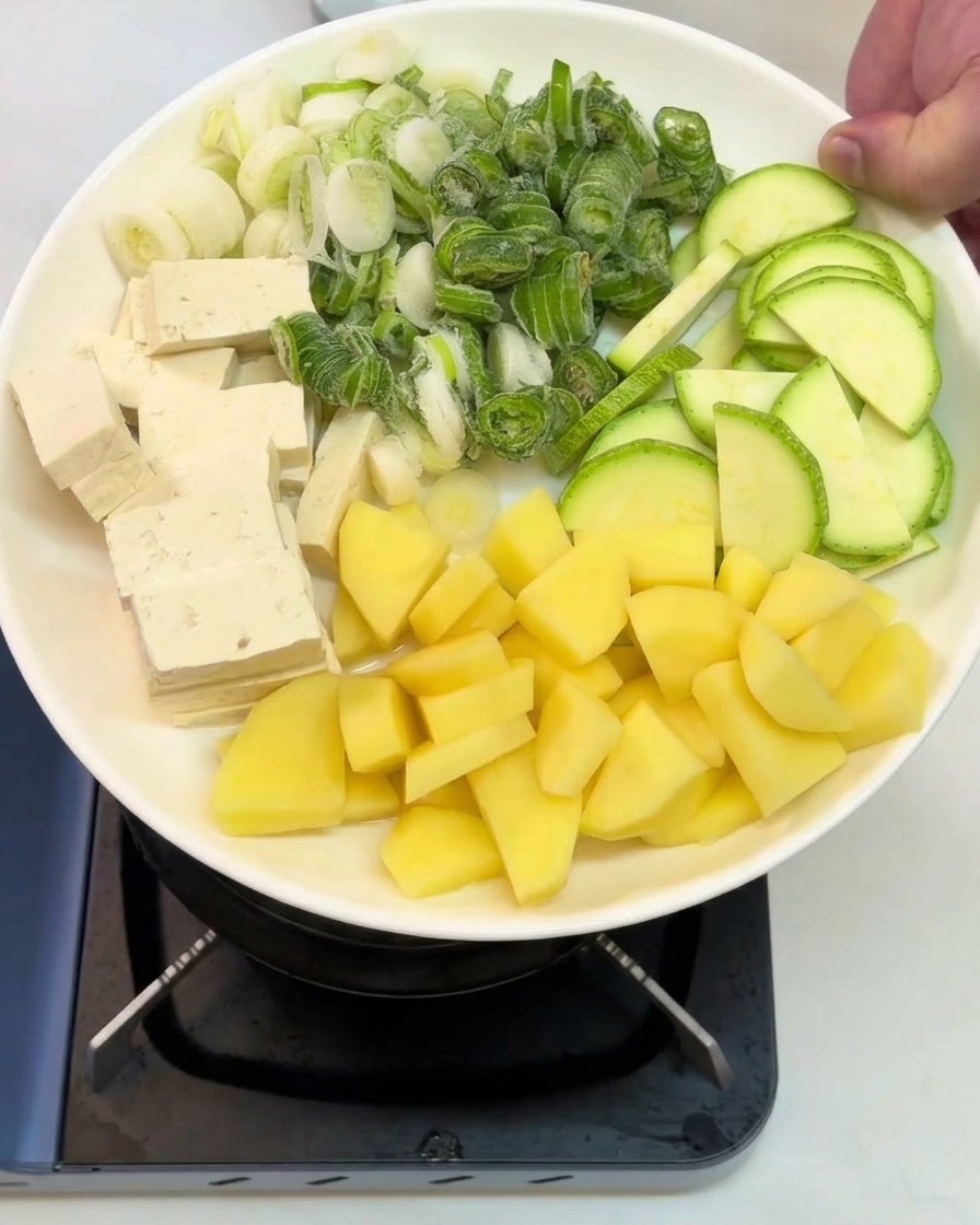 A white plate held over a stove containing neatly organized sections of cubed tofu, chopped green onions, sliced zucchini, and diced raw potatoes.