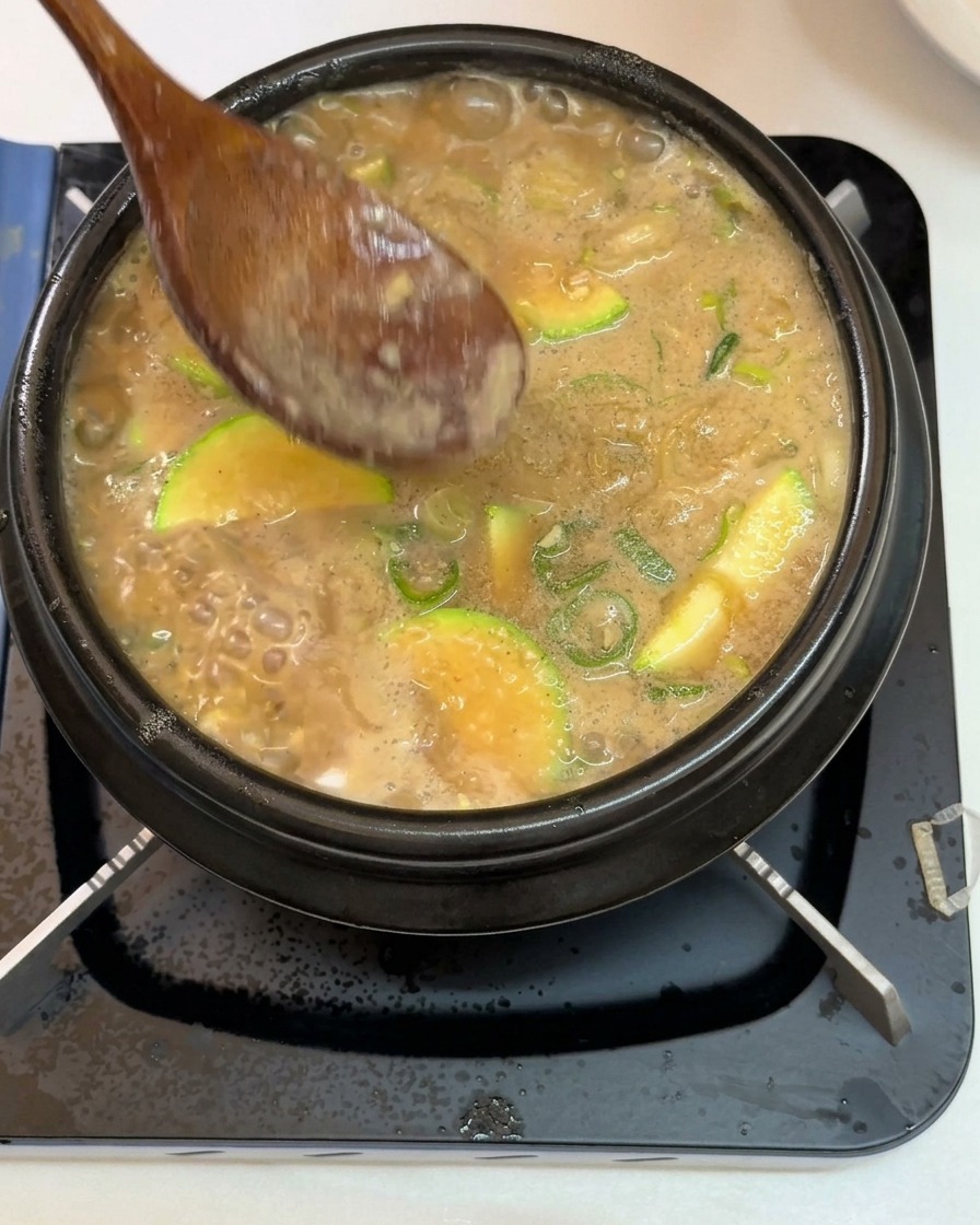 A wooden spoon skimming white foam and impurities from the surface of a bubbling Korean soybean paste stew filled with sliced zucchini and green onions inside a black stone pot.