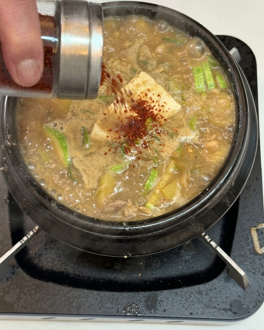 Red chili powder being shaken from a stainless steel and glass jar over a bubbling Korean soybean paste stew filled with tofu and zucchini.
