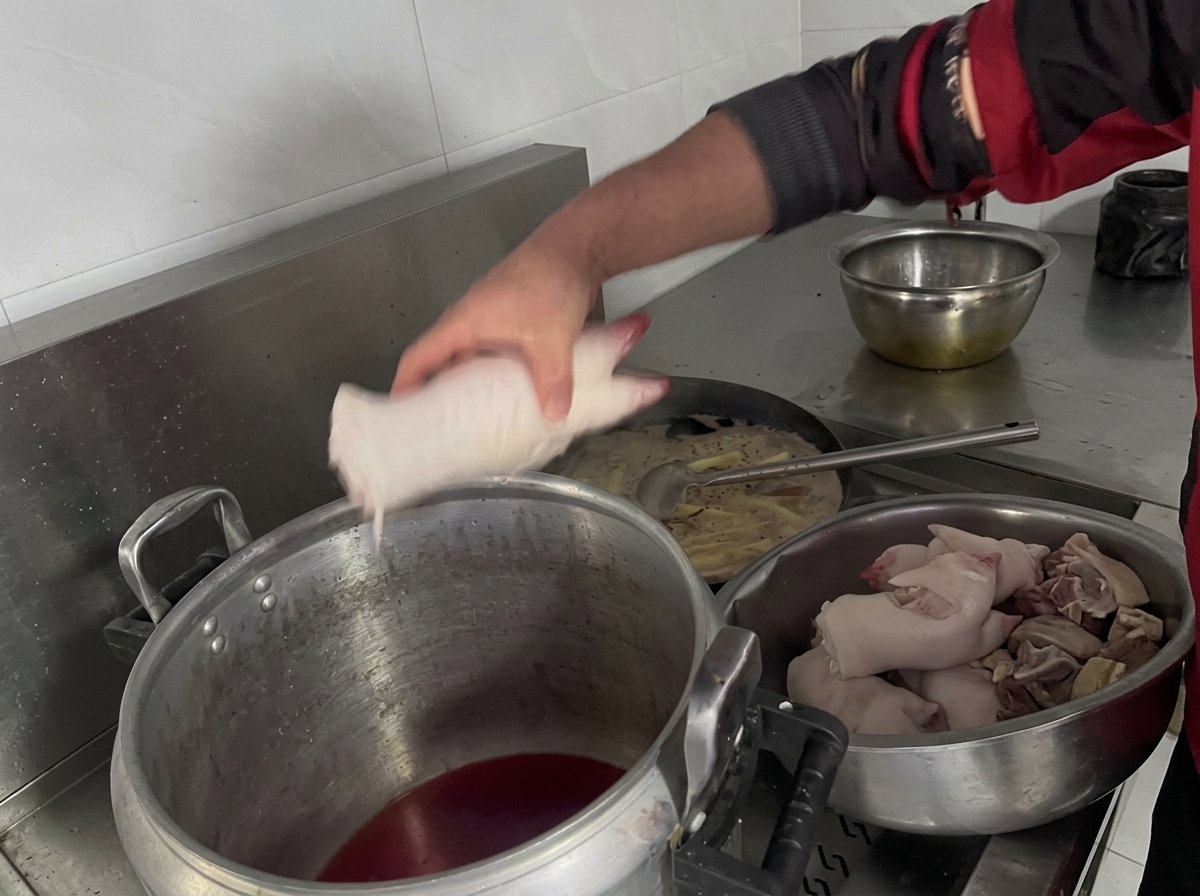 A persons hand placing raw pig trotters into a large aluminum stockpot filled with dark braising liquid.