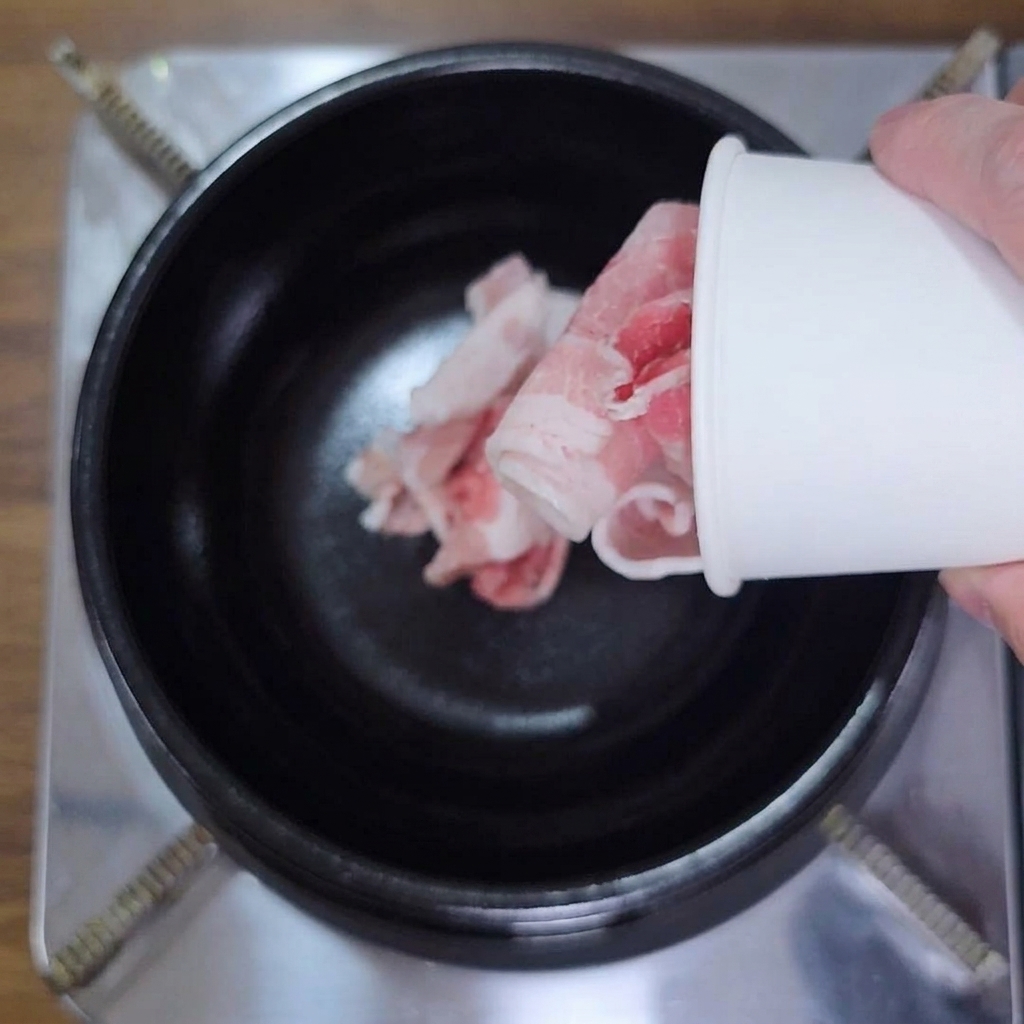 A hand pouring rolled, thinly sliced raw pork belly from a white paper cup into a black pot.