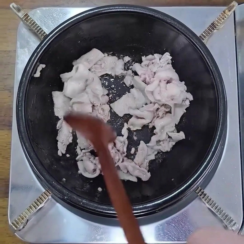 Thinly sliced pork belly being stir-fried in a black pot with a wooden spoon until lightly browned.