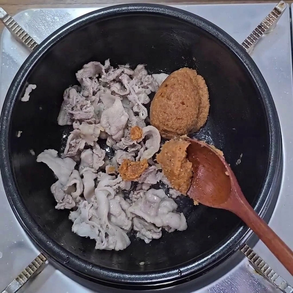 Scoops of brown soybean paste and ssamjang added to cooked pork belly in a pot.