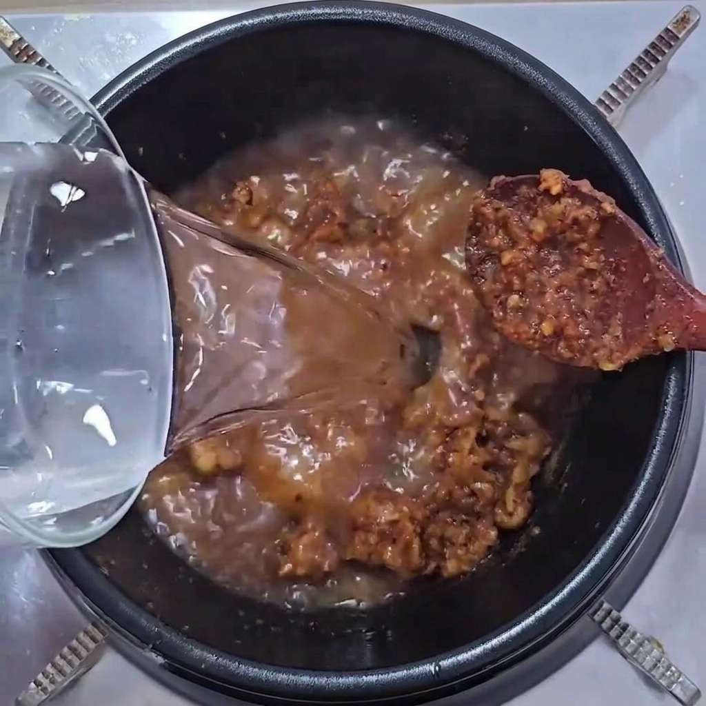 Clear water being poured from a glass container into a pot containing a sizzling mixture of pork and soybean paste.