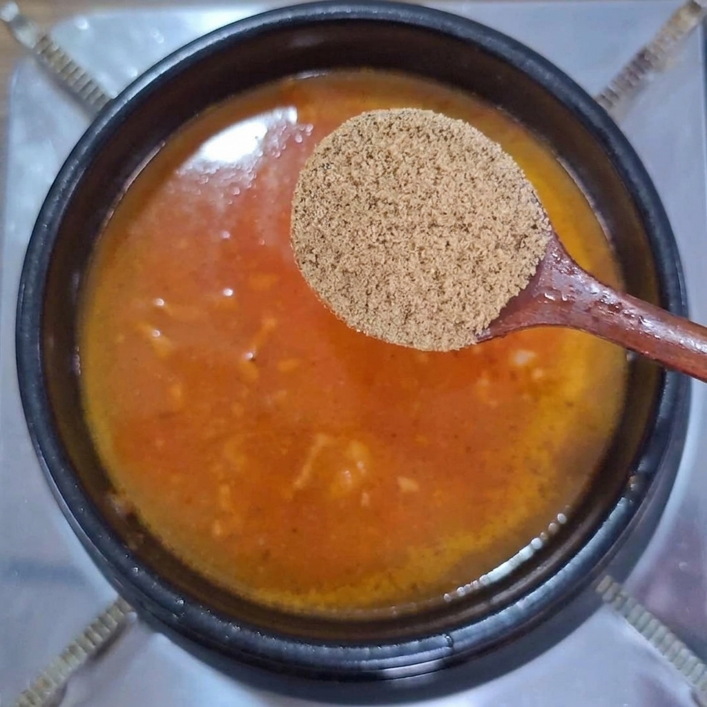 A wooden spoon holding dry beef bouillon powder over a pot of bubbling orange-red broth.