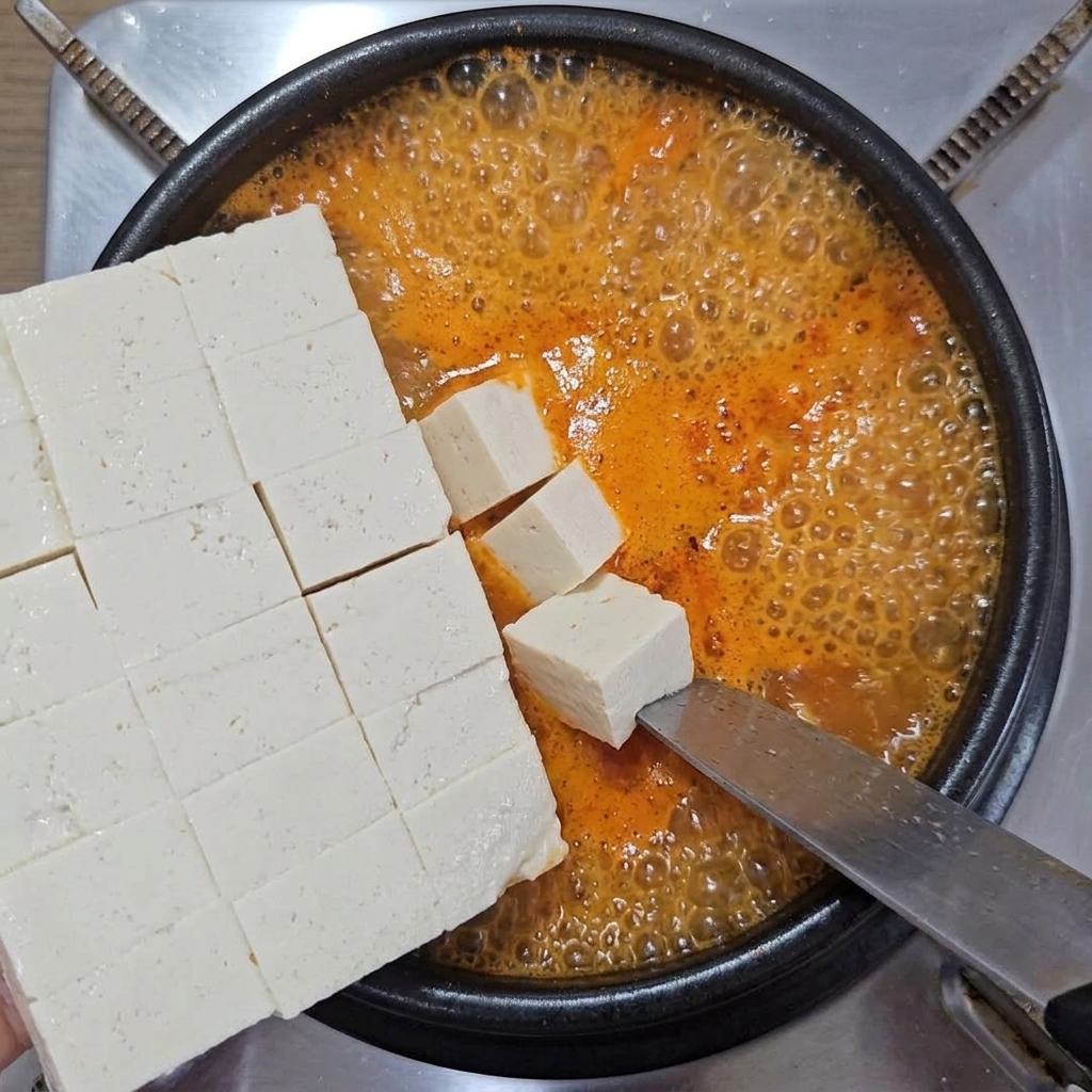 Cubed soft white tofu being gently slid off a large knife into a pot of bubbling orange-red stew.