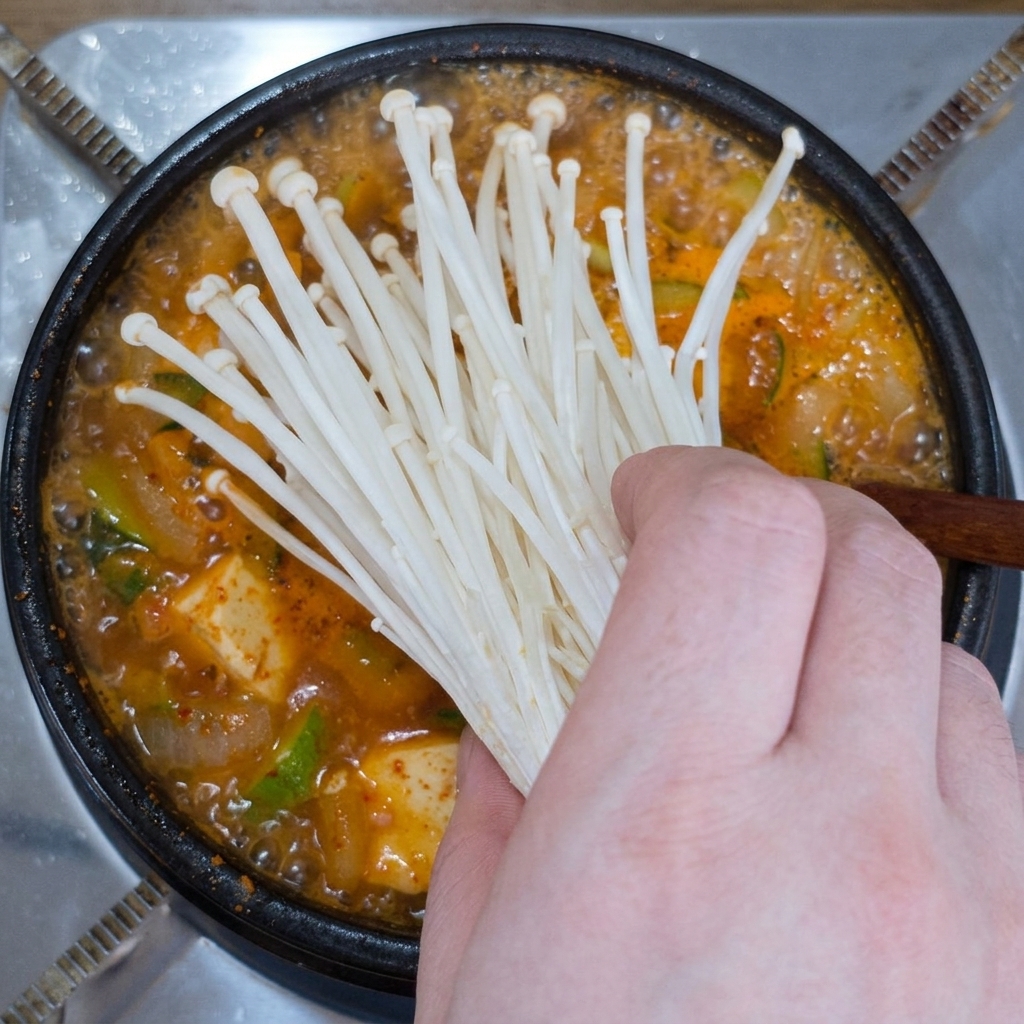 A hand placing a bunch of fresh white enoki mushrooms into a pot of boiling soybean paste stew.