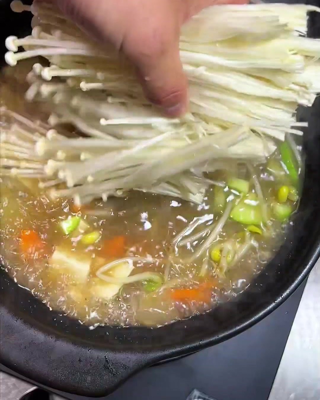 A bundle of delicate, long-stemmed enoki mushrooms being added to the simmering pot.