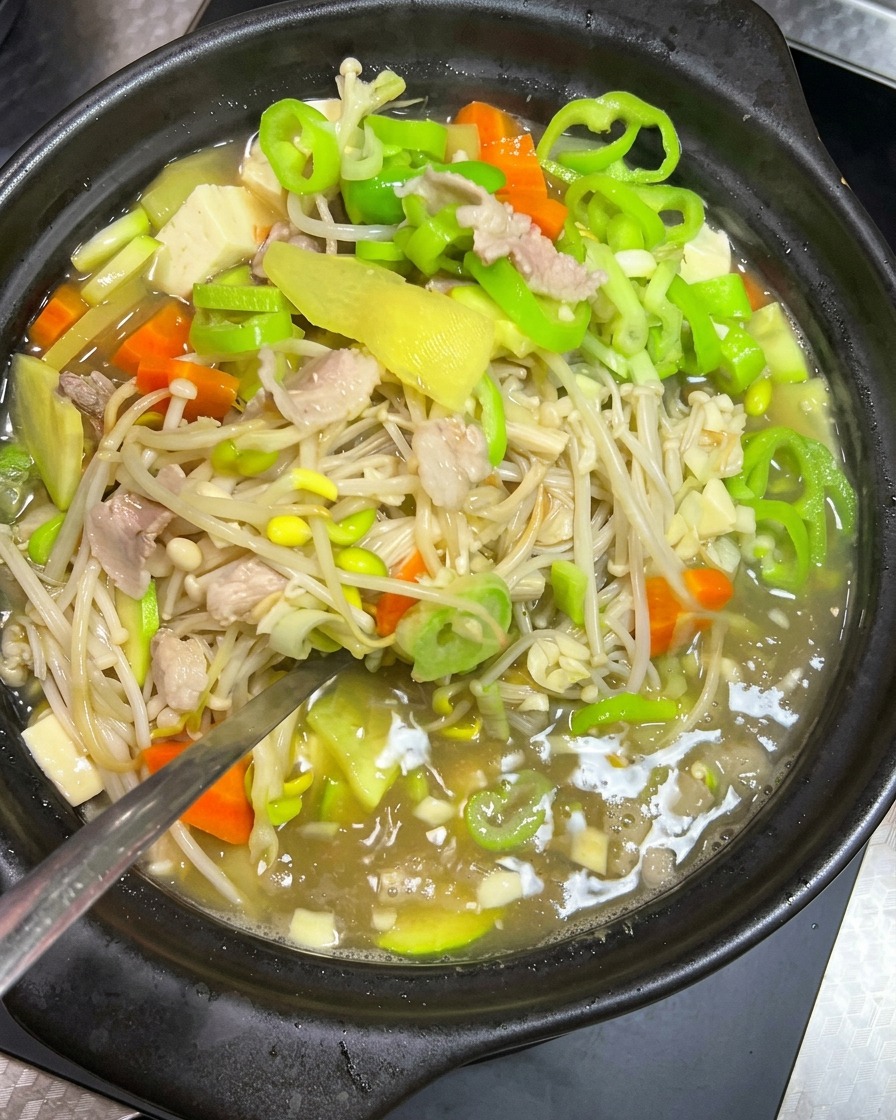 A metal spoon gently stirring a simmering stew of bean sprouts, pork, and green vegetables in a black clay pot.