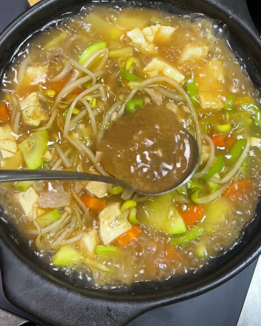 A metal spoon holding dark Korean spicy paste being stirred into a bubbling clay pot filled with tofu, zucchini, bean sprouts, and broth.