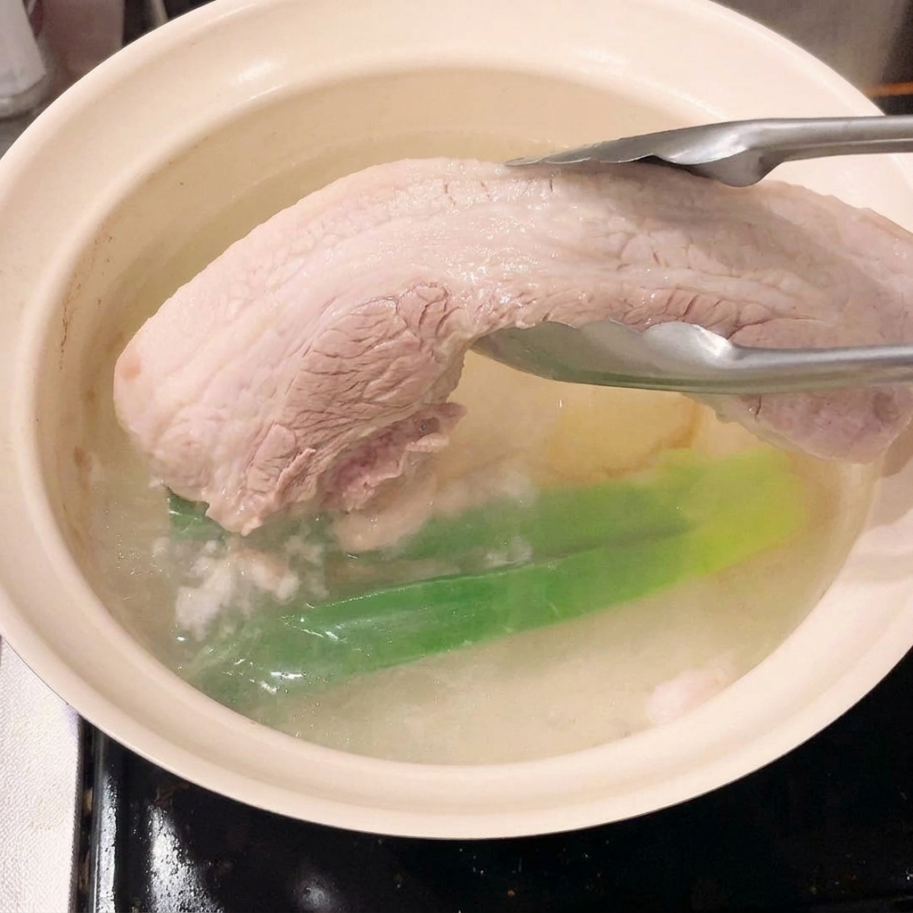Tongs lifting the blanched, firm piece of pork belly out of the cooking water.