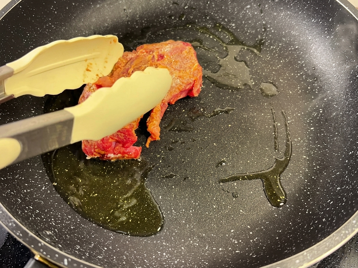 A piece of seasoned beef being seared in a hot pan with oil, tongs holding it in place.