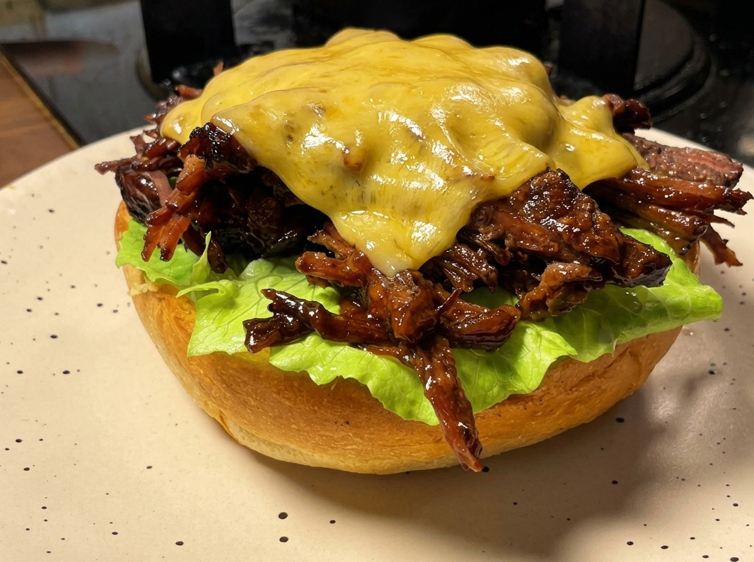 A pulled beef burger patty topped with a cheese slice that is being melted by a handheld kitchen torch.