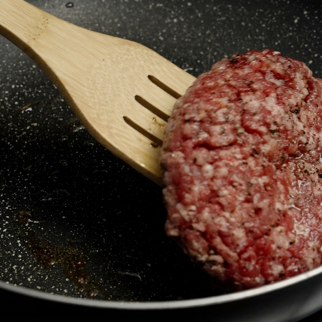A wooden spatula lifting and flipping a thick burger patty that has a beautifully browned, crusty bottom in a hot pan.