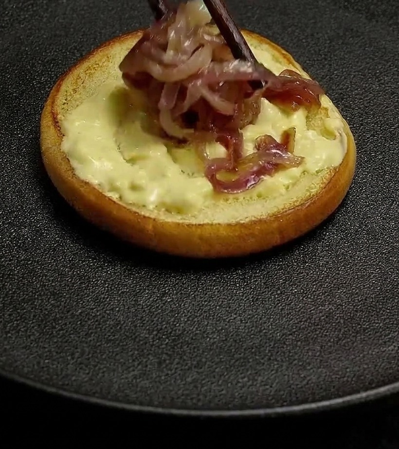 Cooked red onions being placed with chopsticks onto a bun spread with creamy burger sauce.