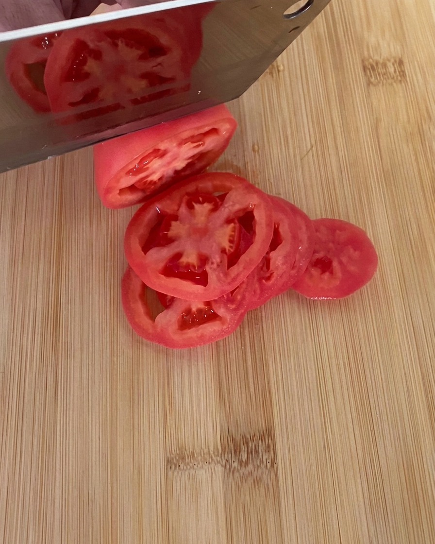 A knife slicing a ripe red tomato into thick rounds on a wooden cutting board.