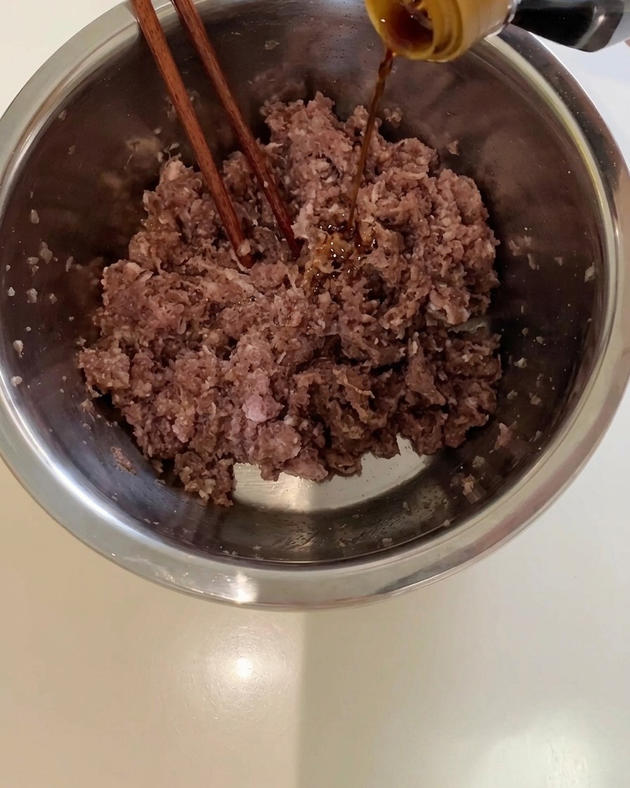 Dark soy sauce being poured from a bottle into a metal bowl filled with ground beef.
