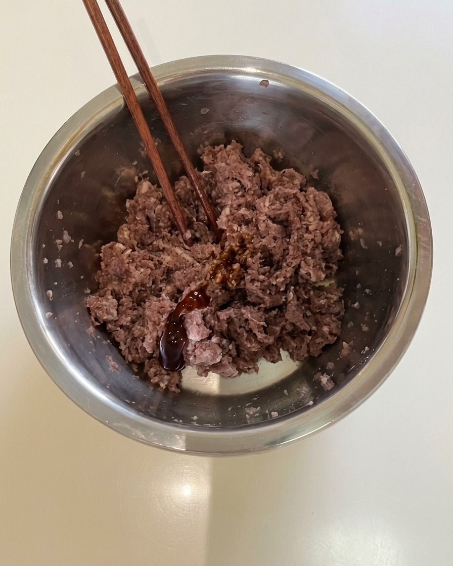 Chopsticks resting in a metal mixing bowl filled with seasoned ground beef and dark sauce.