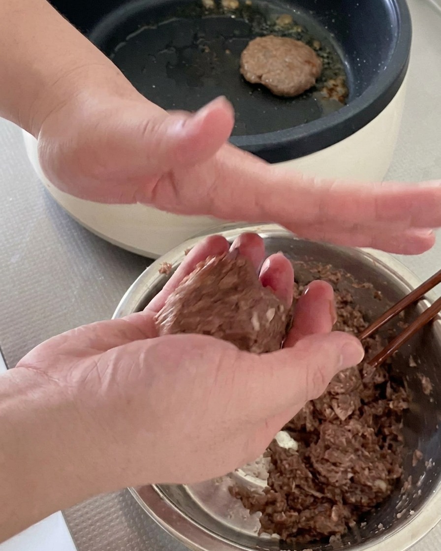 Two hands tossing and shaping a ball of raw ground beef into a round burger patty.