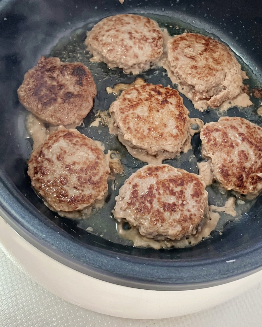 Six round beef patties sizzling and browning in a lightly oiled pan.