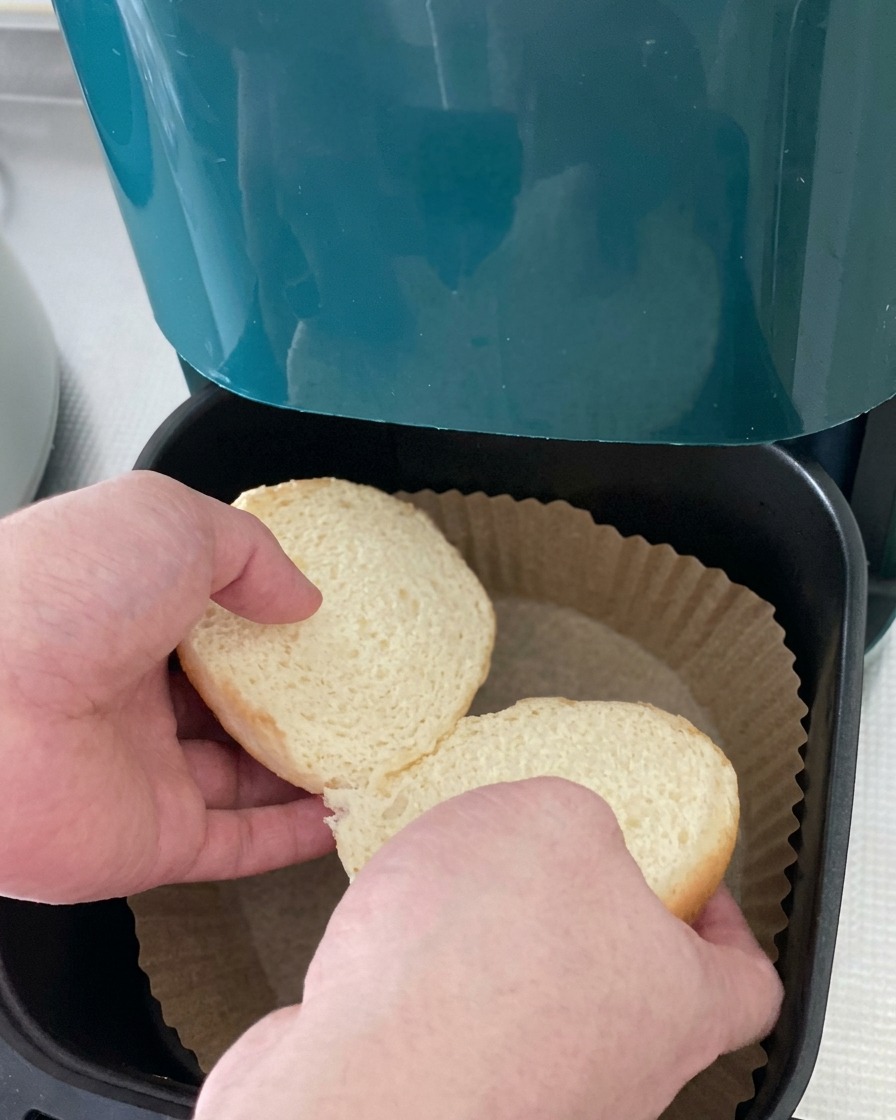 Two halves of a white burger bun being placed into a parchment-lined air fryer basket.