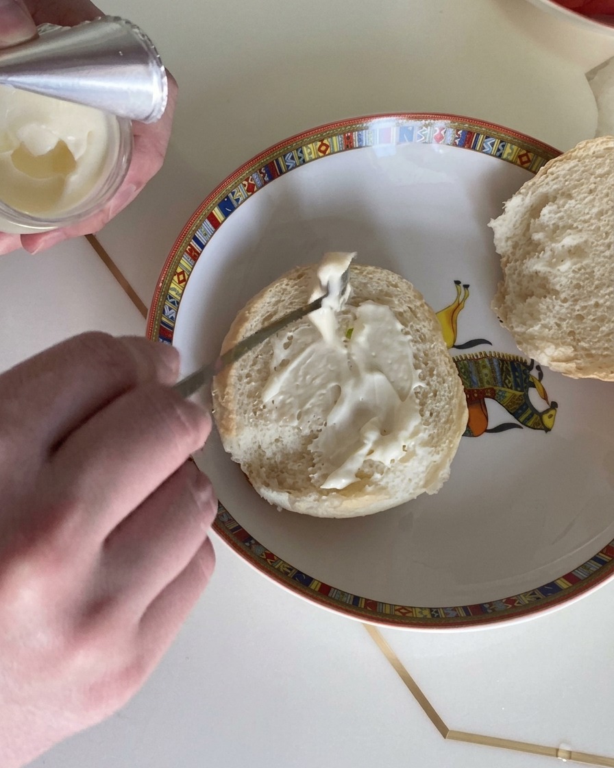 A close-up of a spoon spreading creamy salad dressing onto the cut face of a fluffy burger bun.