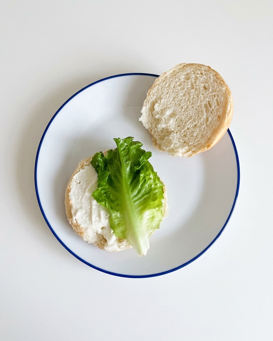 Fresh green lettuce being laid down onto the sauce-covered bottom half of a hamburger bun.