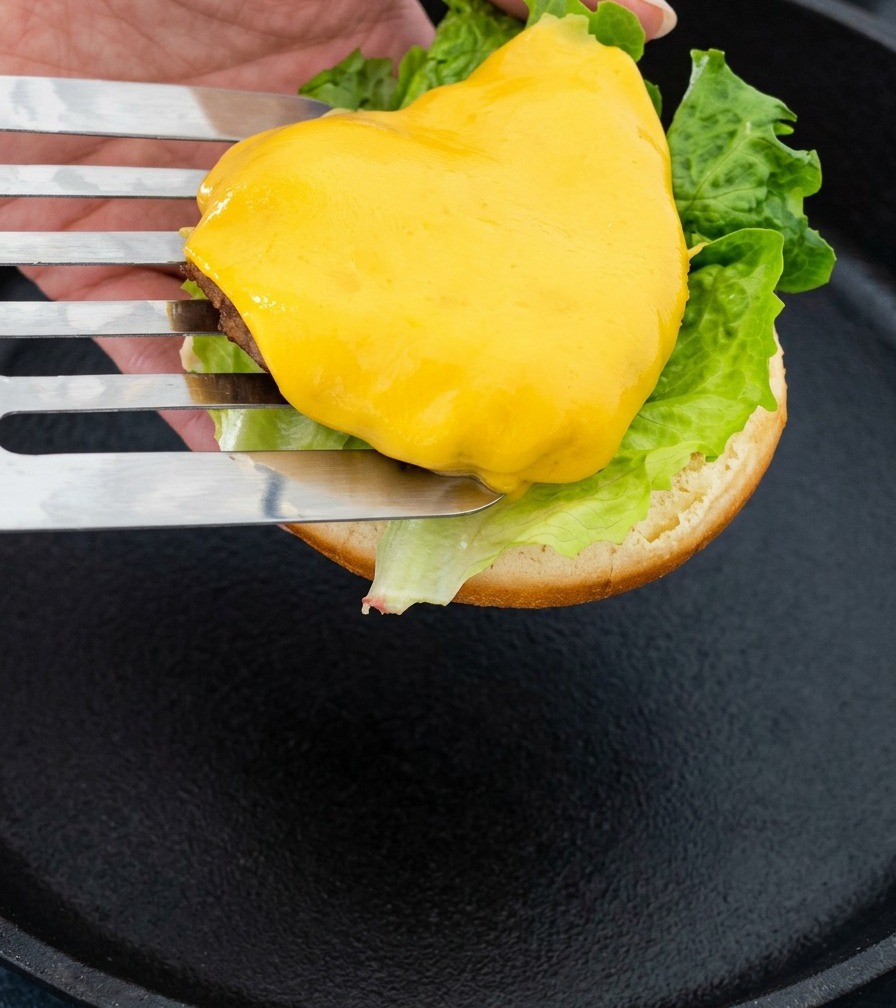 A metal spatula lifting a cheesy beef patty to place it onto a burger bun assembly.