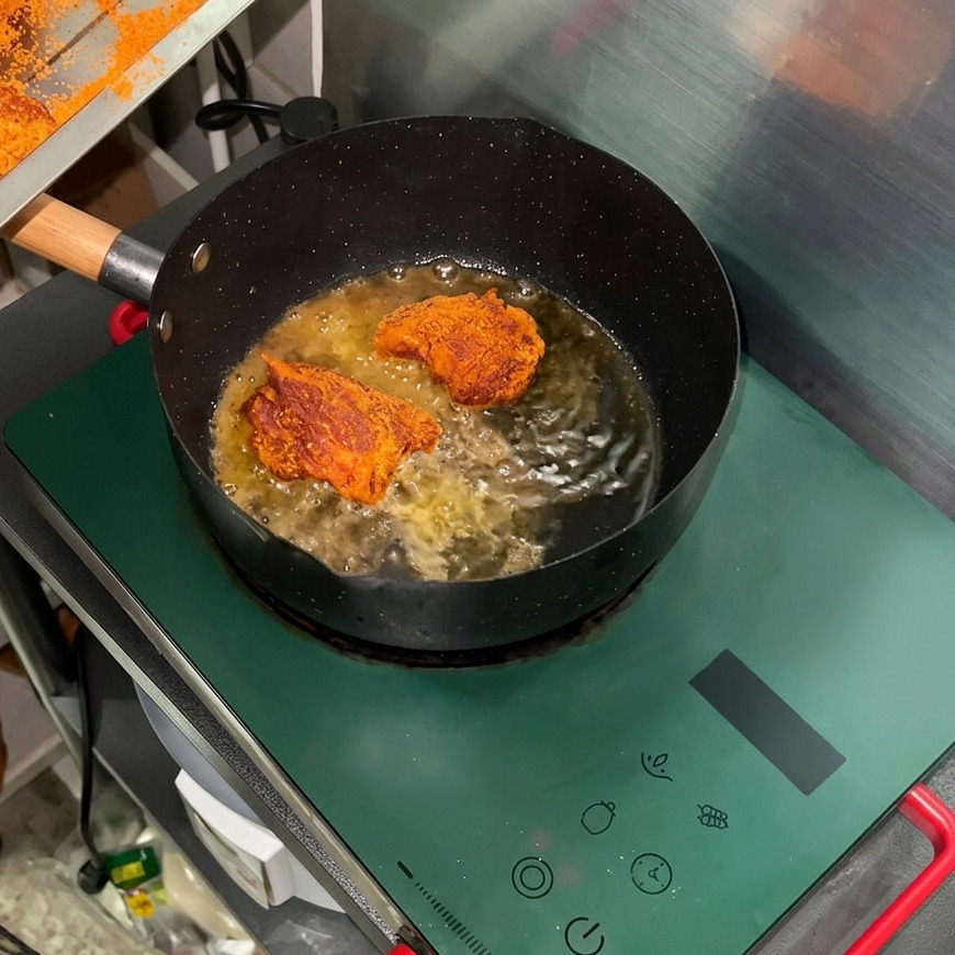 Seasoned pork chunks pan-frying in hot oil inside a black skillet on a green induction stove.