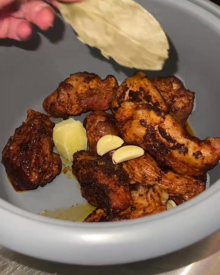 A close-up of a rice cooker pot containing seared pork chunks, fresh garlic, lime pulp, and a bay leaf being added.