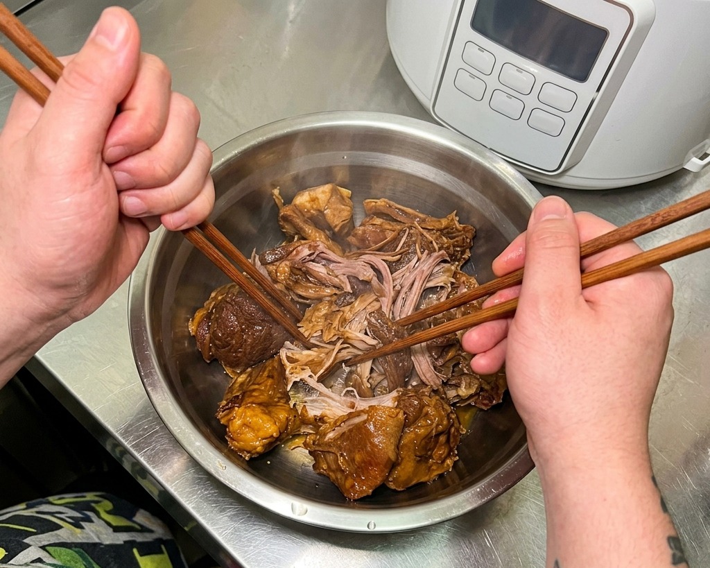 Hands using wooden chopsticks to pull apart chunks of tender, braised pork inside a stainless steel mixing bowl.