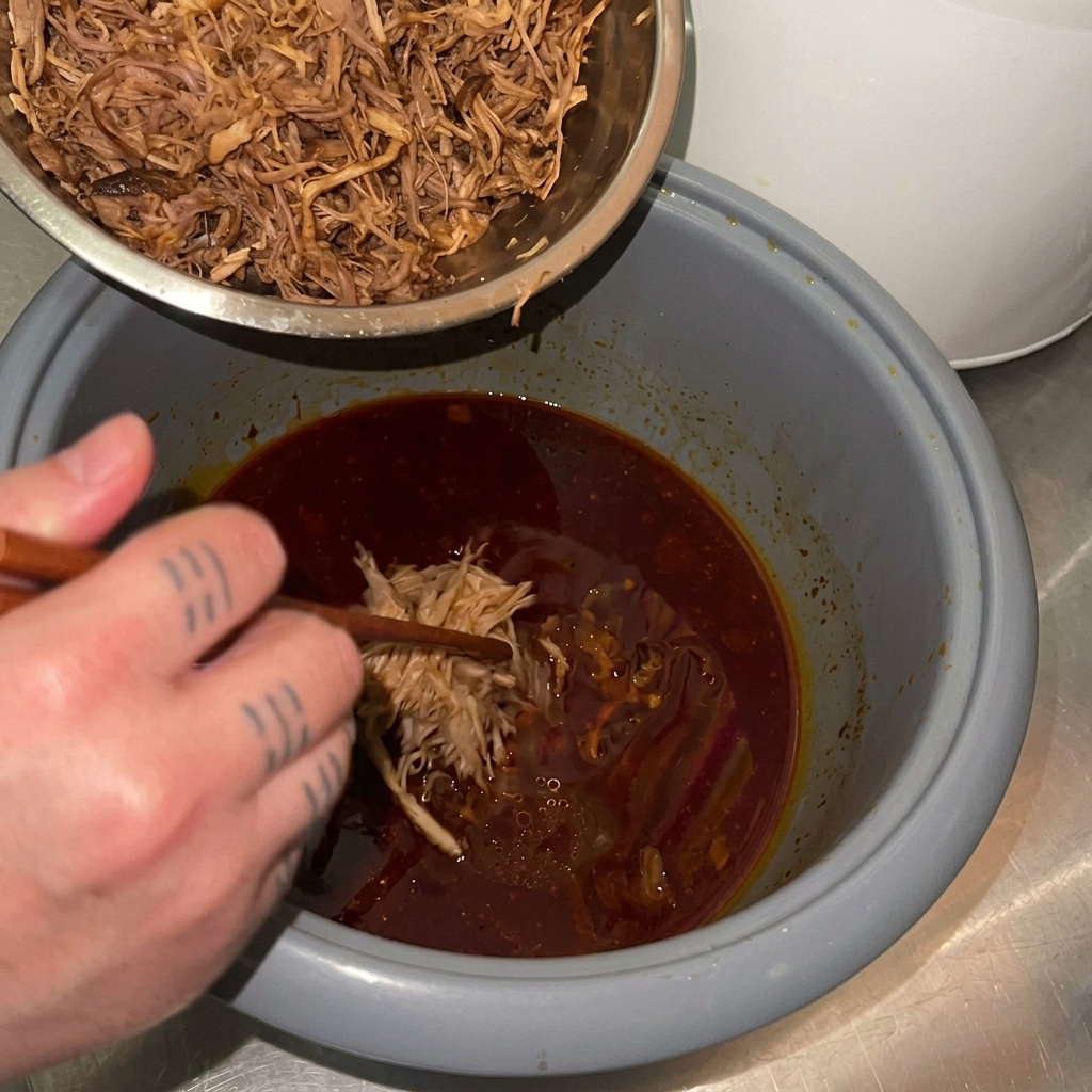 Shredded pork being transferred from a metal bowl back into a rice cooker pot containing a dark red braising sauce.