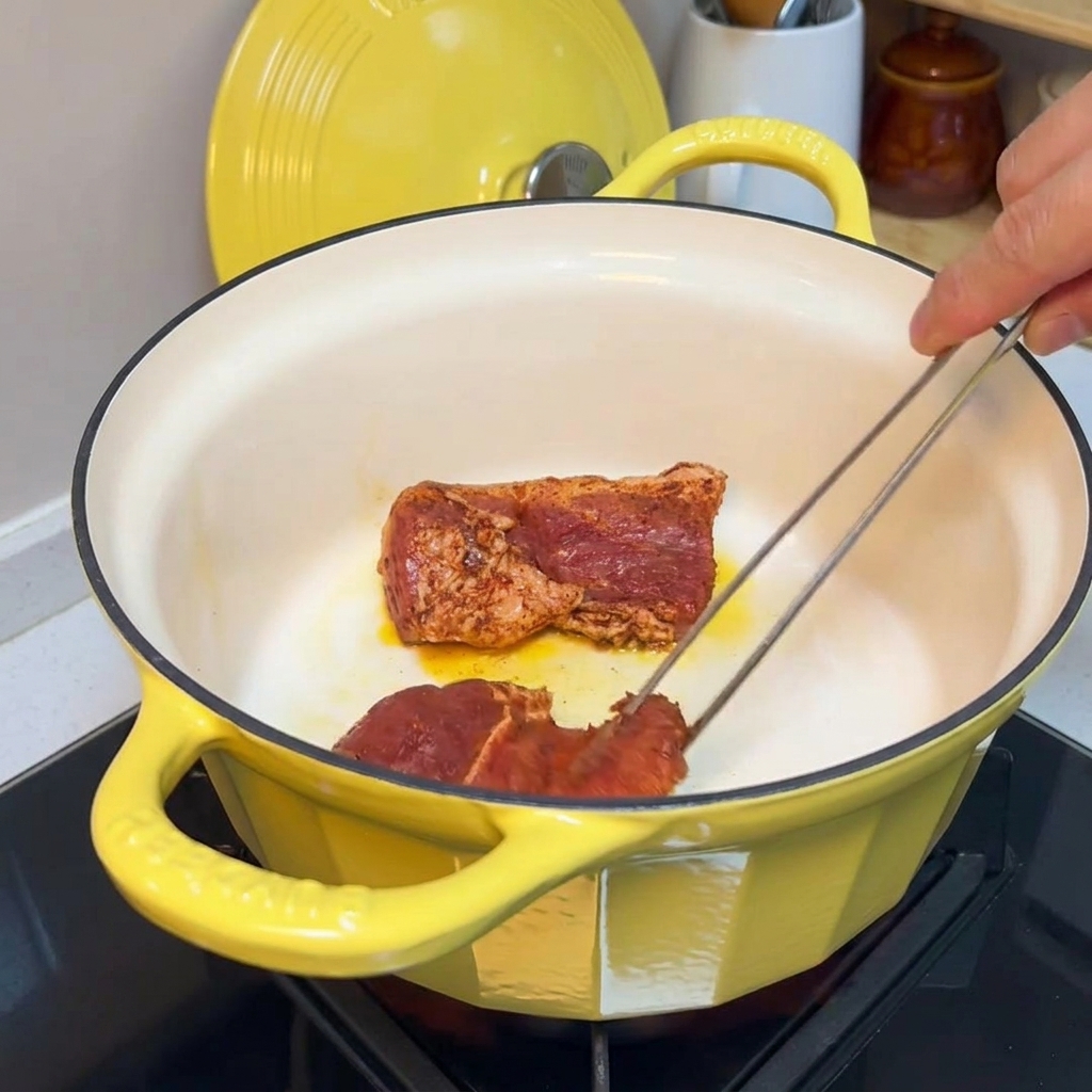 Tongs turning seasoned pork chunks in a yellow cast iron pot to sear the edges.
