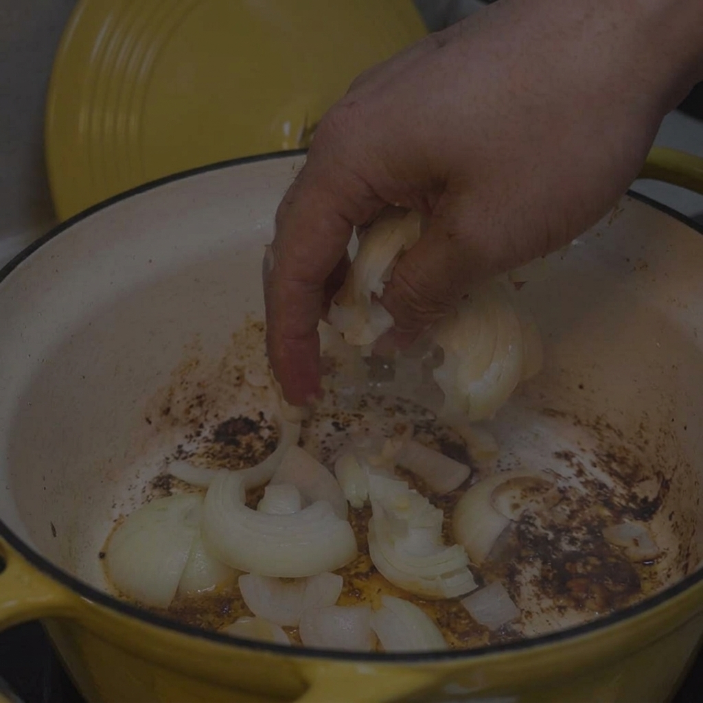 A hand dropping sliced white onions into a yellow pot with browned bits on the bottom.