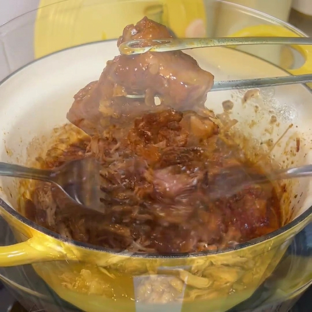 Tender cooked pork shoulder being shredded apart with two forks in a clear glass mixing bowl.
