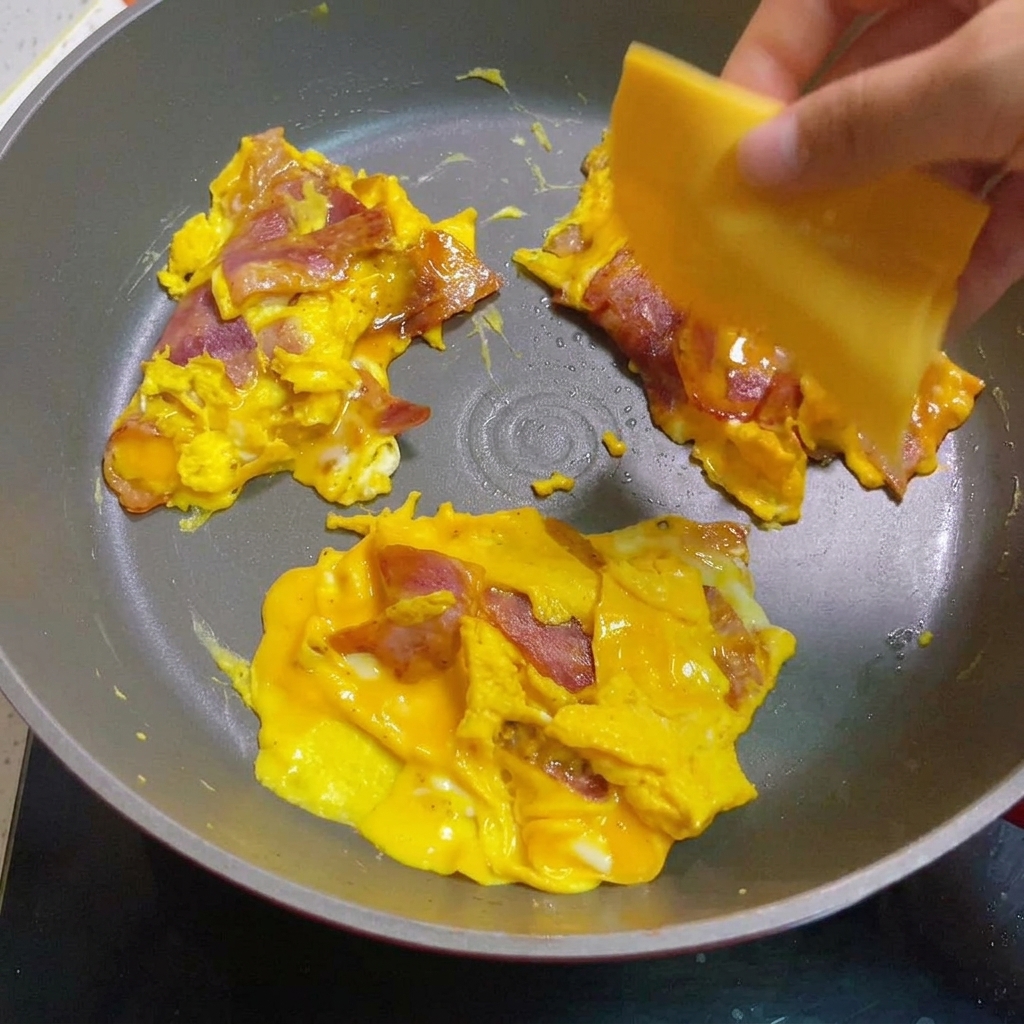 Scrambled eggs and bacon topped with melting cheese slices in a pan.