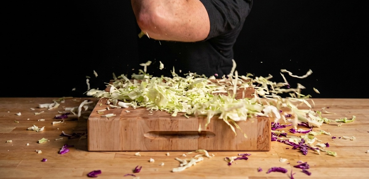 Fresh green and purple cabbage being aggressively prepped and shredded on a large wooden cutting board.
